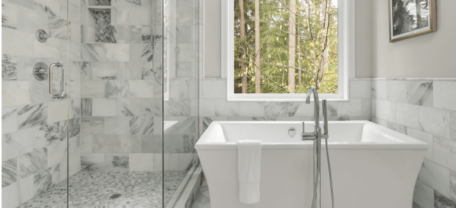 Bathroom with a marble-tiled shower, freestanding bathtub, and large window overlooking trees. A towel hangs over the tub's edge.