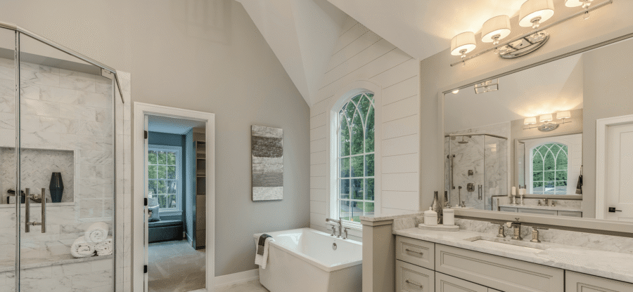A modern bathroom with a large mirror, double sink, bathtub, and glass shower. The room has a vaulted ceiling and a large window with outdoor views.